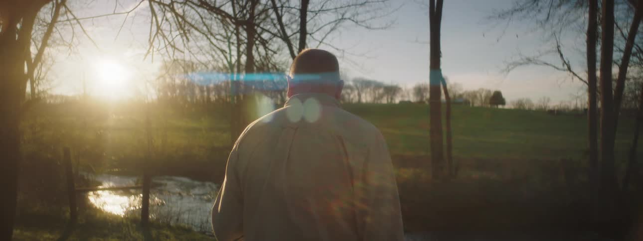 A man stands in a rural grass area beside a small pond