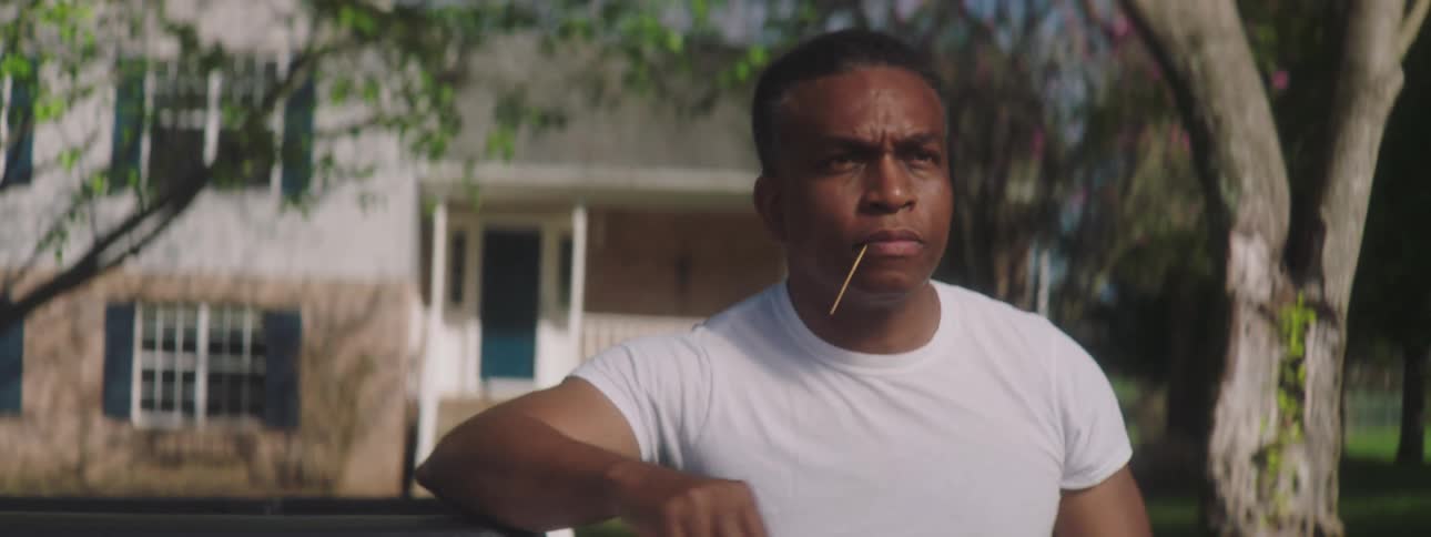 A man wearing a white tee shirt and leaning on the back of his truck sticks a straw in his mouth to chew on