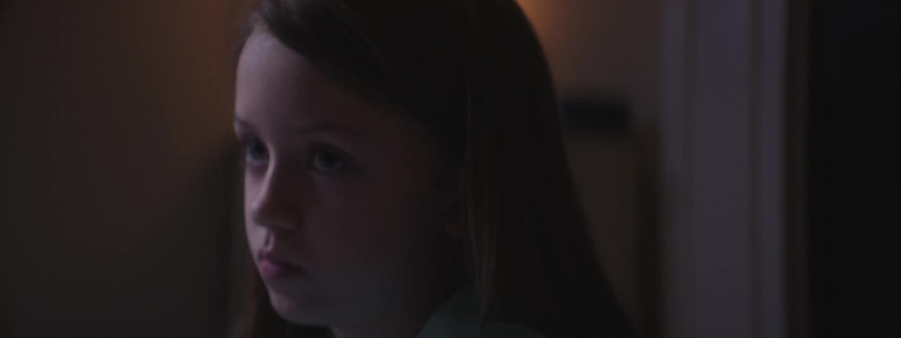 A girl sits in a dim room drawing on white paper with crayons