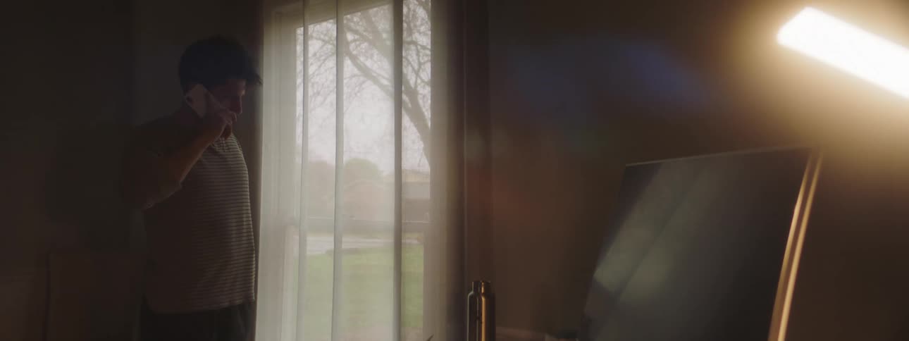 A man standing near a window in his house talks nervously on his smartphone