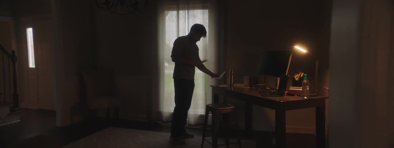A man standing near his desk in a dimly lit room puts things on and takes things off his desk