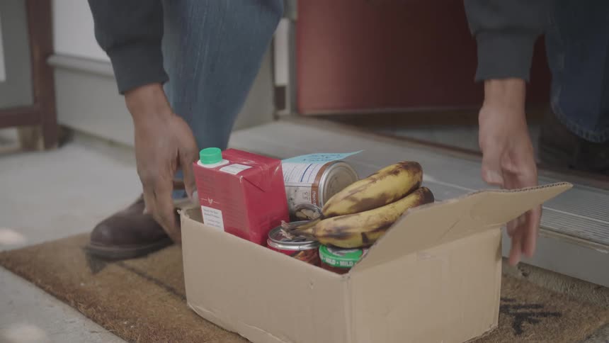 A man opens his door and picks up a box of groceries left on his doorstep