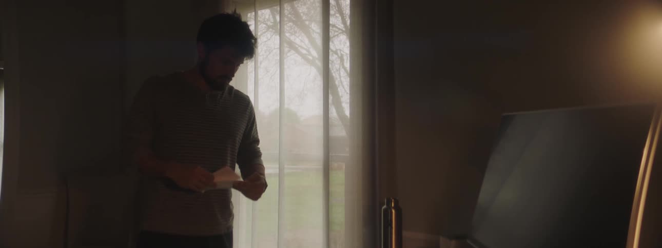 Bearded young man opens mail in a dim lit room