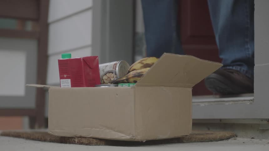 A person leans down to pick up a box of food on their doorstep then goes back inside
