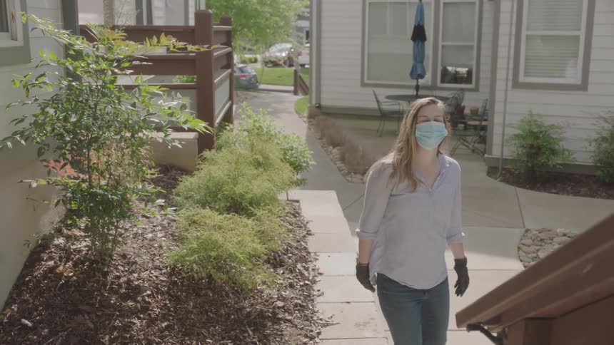 A woman in a face mask descending a staircase from a house and waving back before walking through the yard towards the road