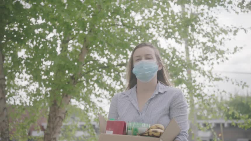 Woman wearing face mask and gloves carries groceries in a box