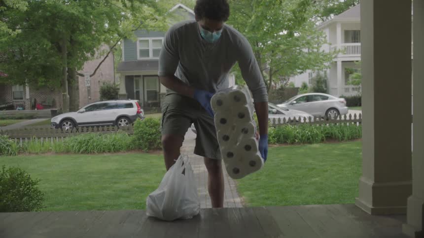 A man carrying a bag and papergoods walks through the yard and up the steps where he leaves the packages and waves