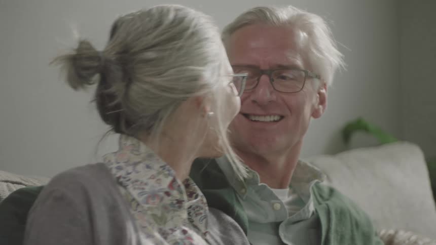 Elderly man and woman sitting on the sofa looks at each other and smiles happily in the living room