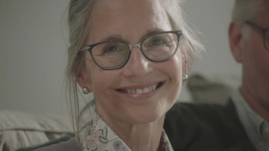 An elderly woman sitting on a sofa raises her head to smile at the camera