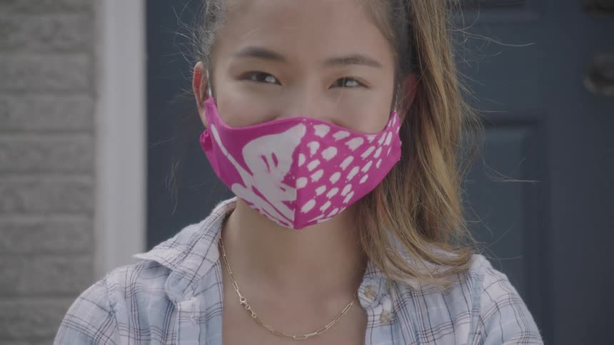 A woman wearing a protective mask and gloves sits on her front stoop holding up a yellow paper heart