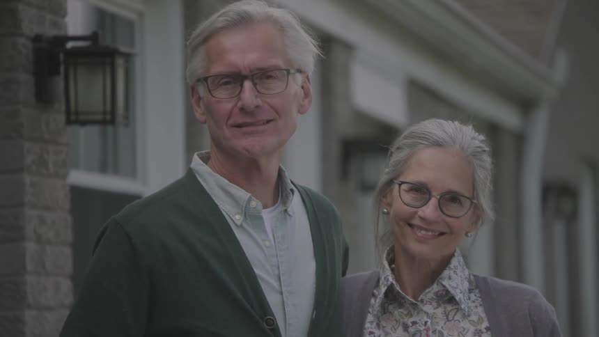Elderly couple stands outside house smiling
