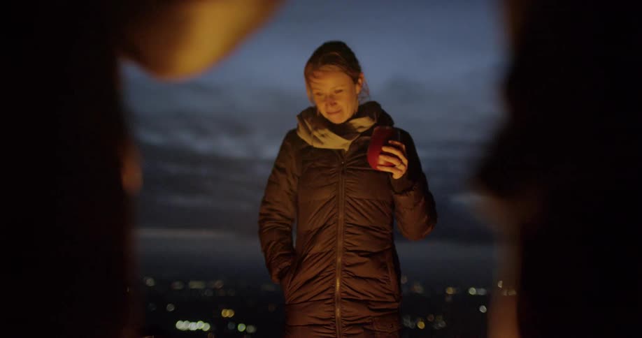 A woman stands at a campfire with a drink in her hand