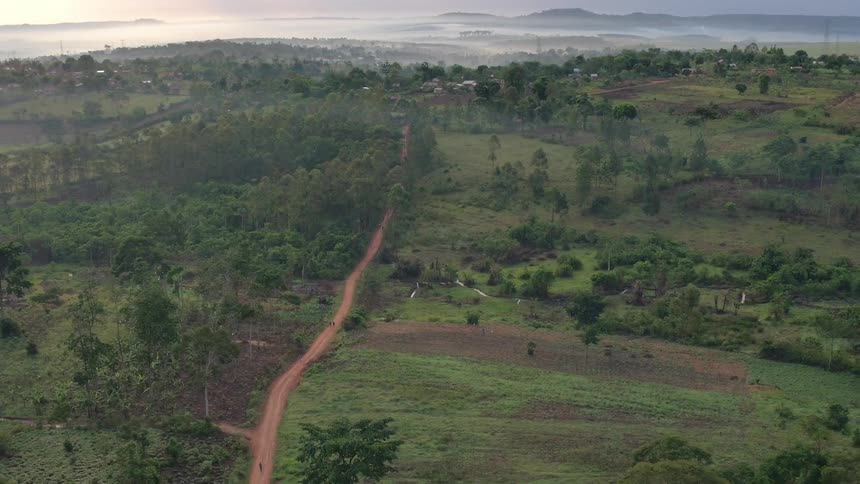 Sun turns fog on horizon red as dirt road leads toward fog shrouded rural Uganda, Africa