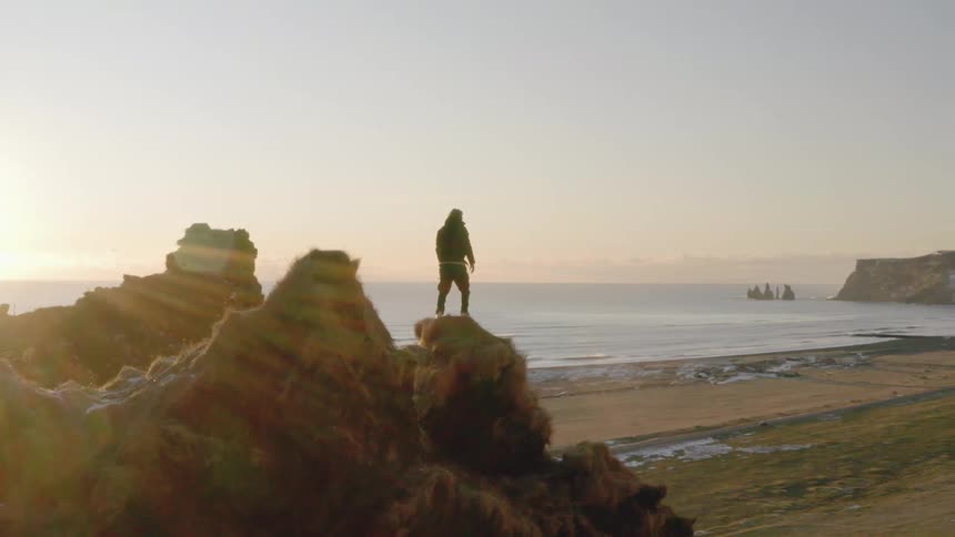 Man stands on top of rocky cliff looking at sunset next to the ocean