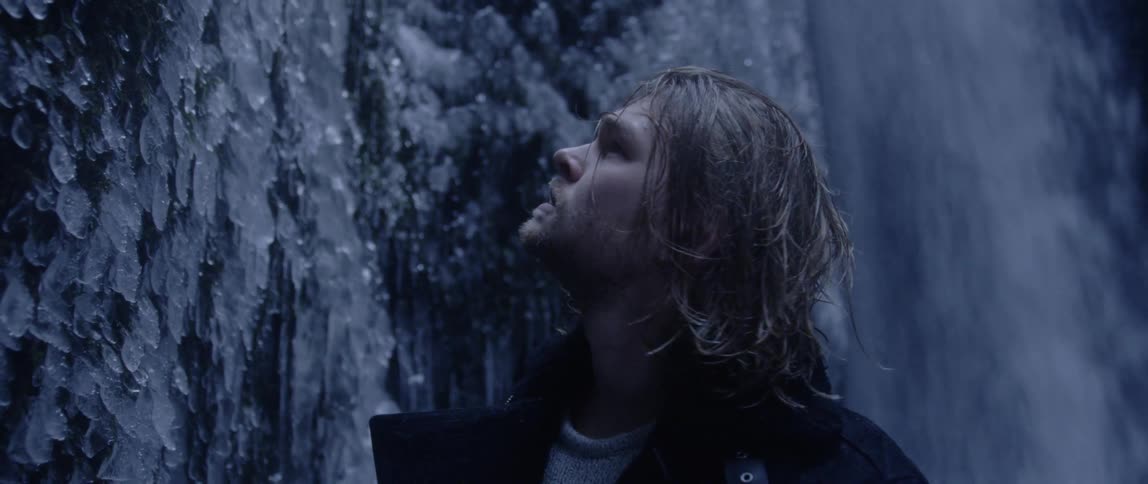 Man wearing coat stands near waterfall looking at icicles