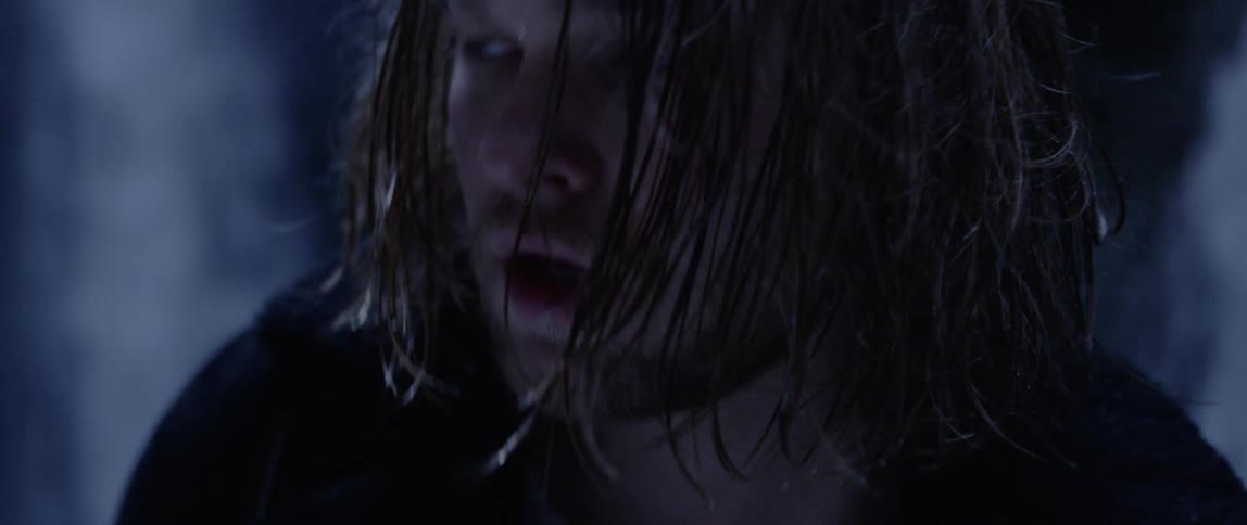A man with damp hair looks up while water from a waterfall flows down rock walls behind him