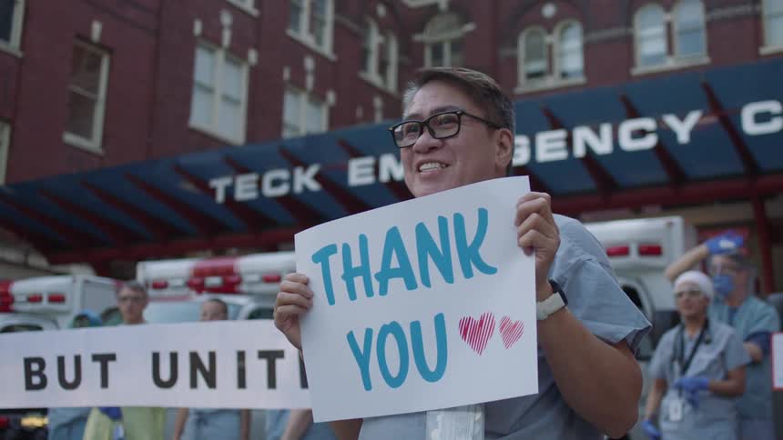 Health care workers stand outside a hospital and wave and hold thank you signs