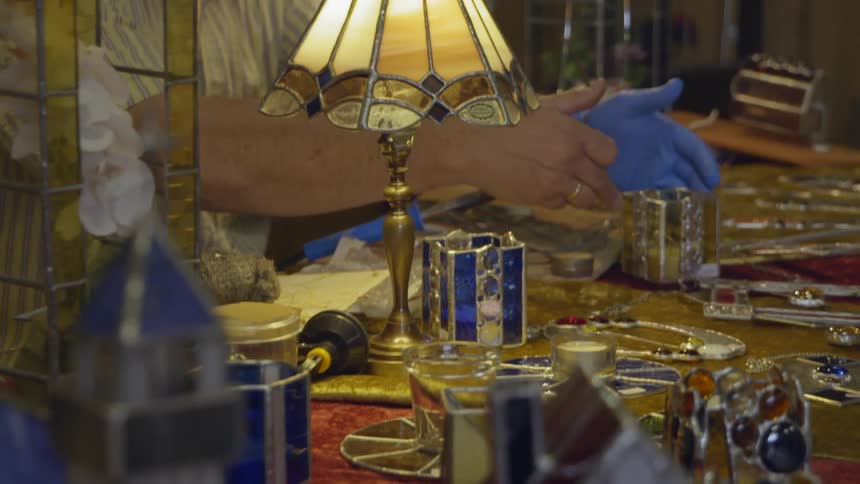 Person stands behind counter with stained glass items gesturing with a glove on one hand