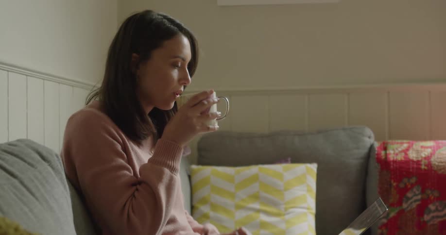 A woman drinks tea while using a laptop on a gray couch with throw pillows