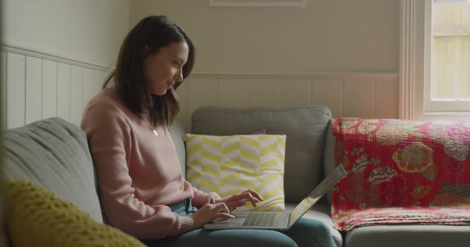 A woman smiles while typing on her laptop while sitting on the couch in her living room
