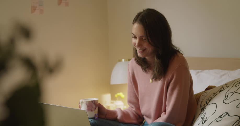 A woman laughing while sitting on a bed looking at the screen of a laptop computer