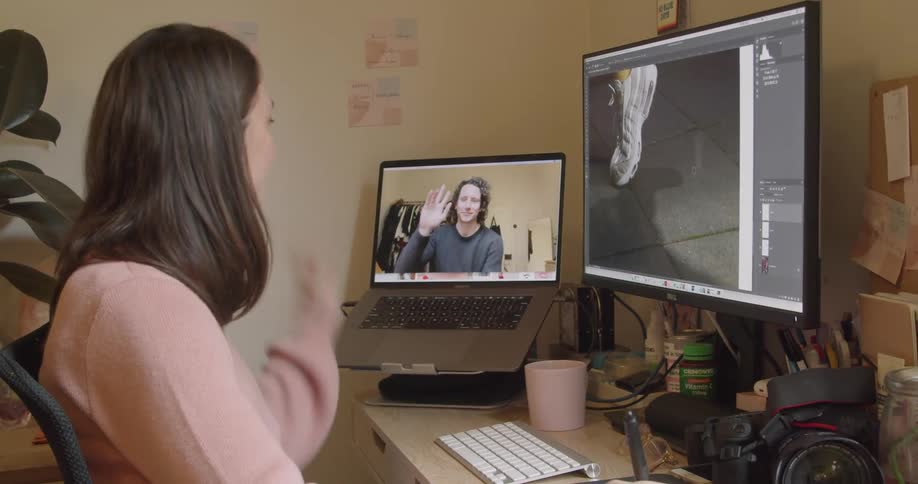 A white woman waves to a man through a computer camera while sitting at a desk