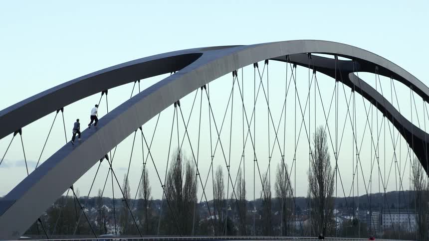 Cars travel across a bridge while two men run up the curved beam on top