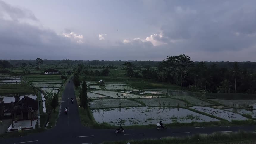 People on the street riding motorcycles with rice patties and the sky in the background