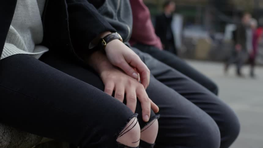 A man and woman sit together and rub hands as people walk by in the distance