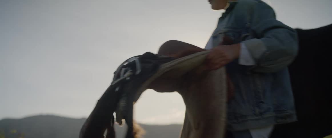 A woman takes a saddle off a horse in a field and carries it away leaving the horse to graze