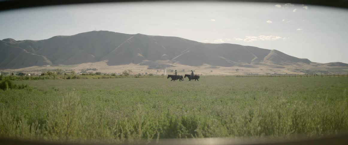 Two people ride horses through the grass of a field with mountains behind on a sunny day