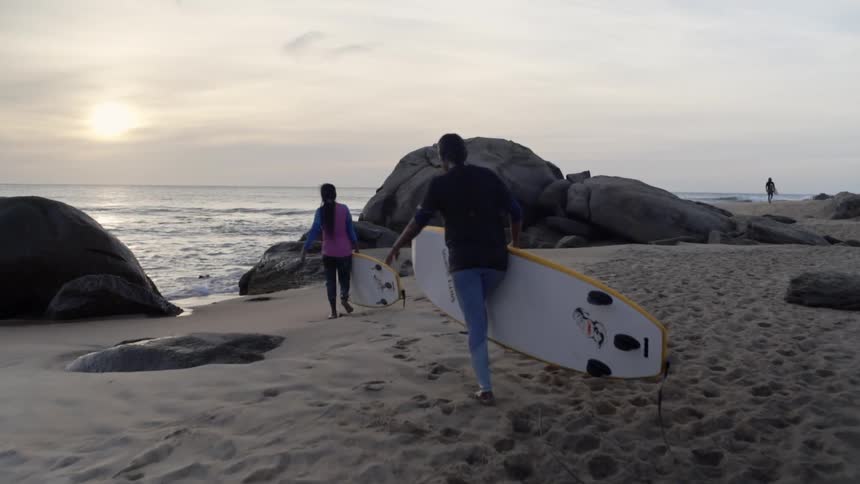 Two surfers carry surfboards on a sandy beach at sunset