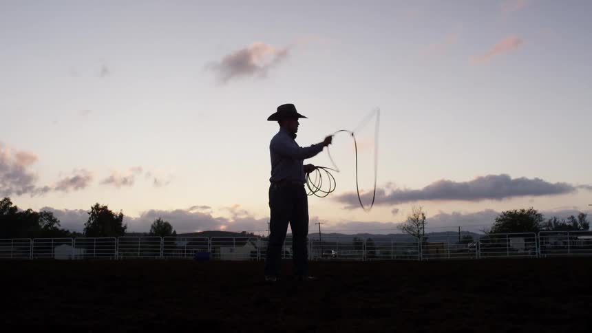A cowboy stands in a paddock practicing twirling a lasso at sunset