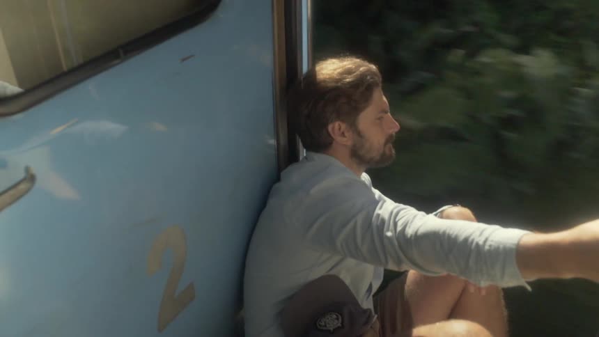 A man sitting on a train with the door open with trees in the background