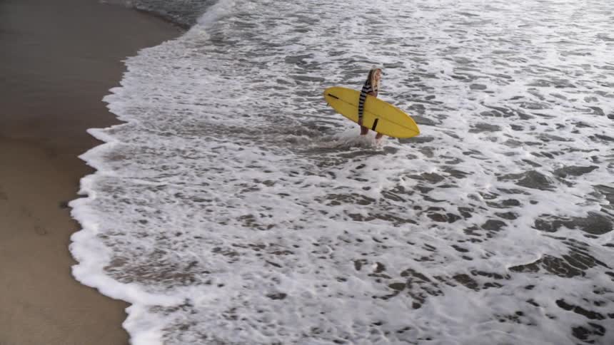 A surfer carrying a yellow board wades into the ocean