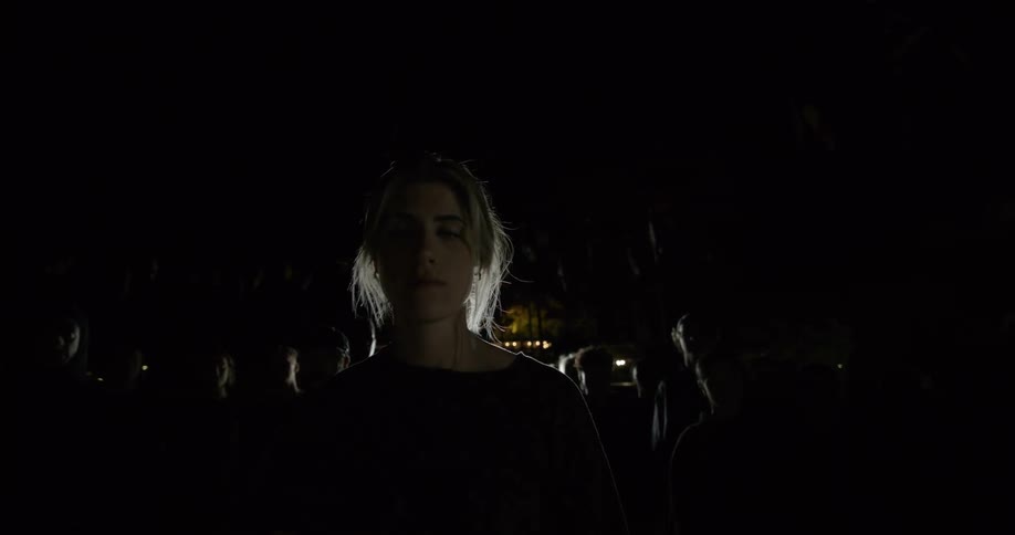 A white woman stands and stares outdoors in front of a large crowd of people