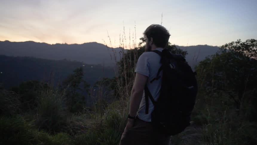 Bearded man carrying backpack hikes throug mountains at sunset