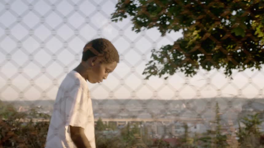 A boy walking with a chain link fence in the foreground and a city skyline in the background
