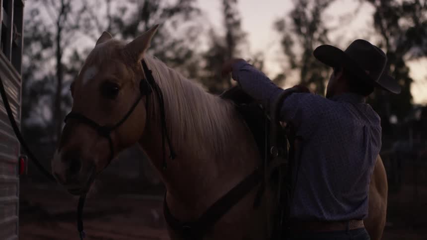 A man with a hat standing next to a horse taking a rope from its saddle