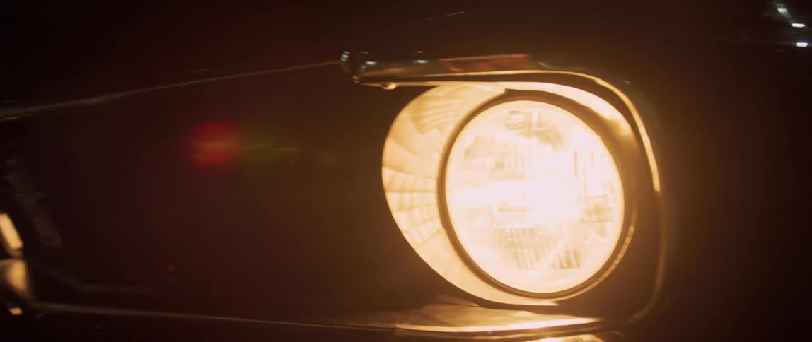 A vehicle headlight is shown up close as the vehicle drives along a road at night