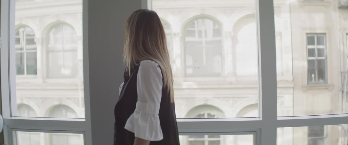 Blonde woman in a dress walks in a room past the windows