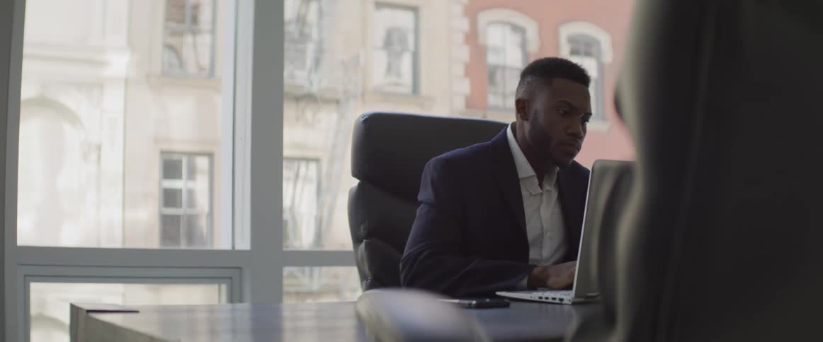 Black man sits at an office desk as he works on a laptop