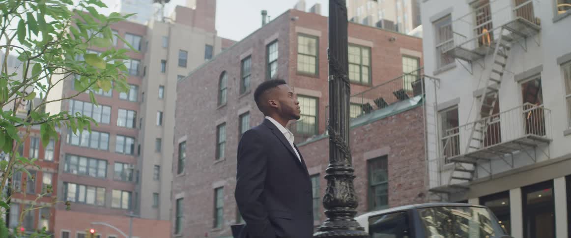 A man in a suit jacket stands next to a city street and looks up