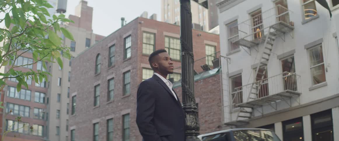 A man wearing a suit coat stands on a city street looking ahead and thinking