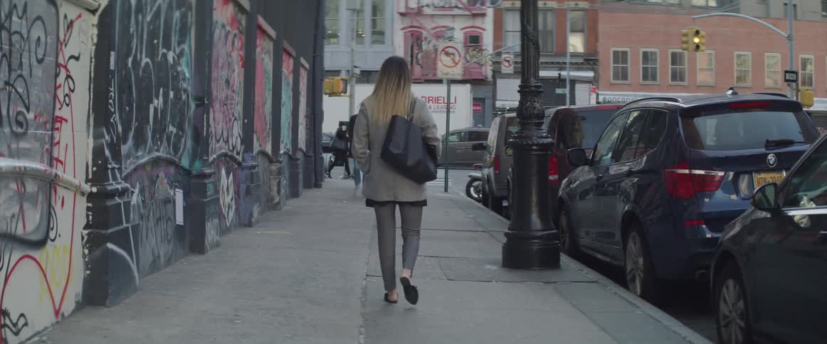 Female walks down a city sidewalk next to a building covered in graffiti on her left and cars parked along the side of the street on her right