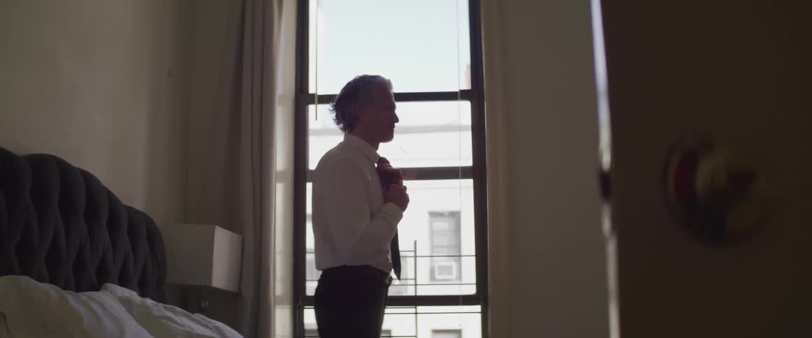 A man stands in front of a mirror putting on a tie likely in a hotel room