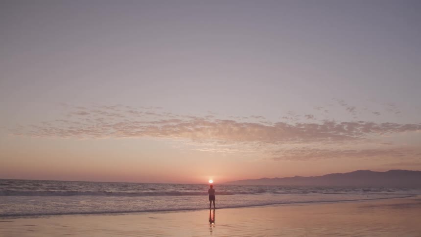 Woman stands on the wet sand beach and watches the sunset