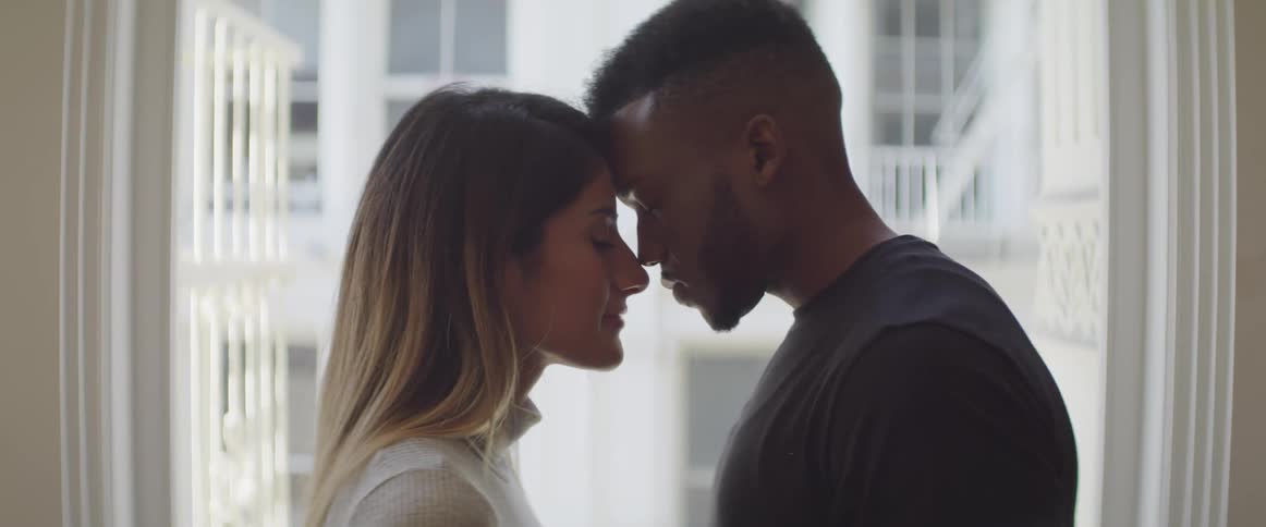 Interracial couple touching foreheads in front of a large window