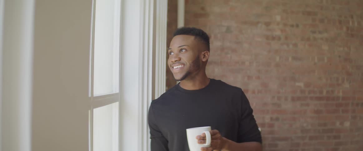 Young black man smiling leaning next to window holding a coffee mug