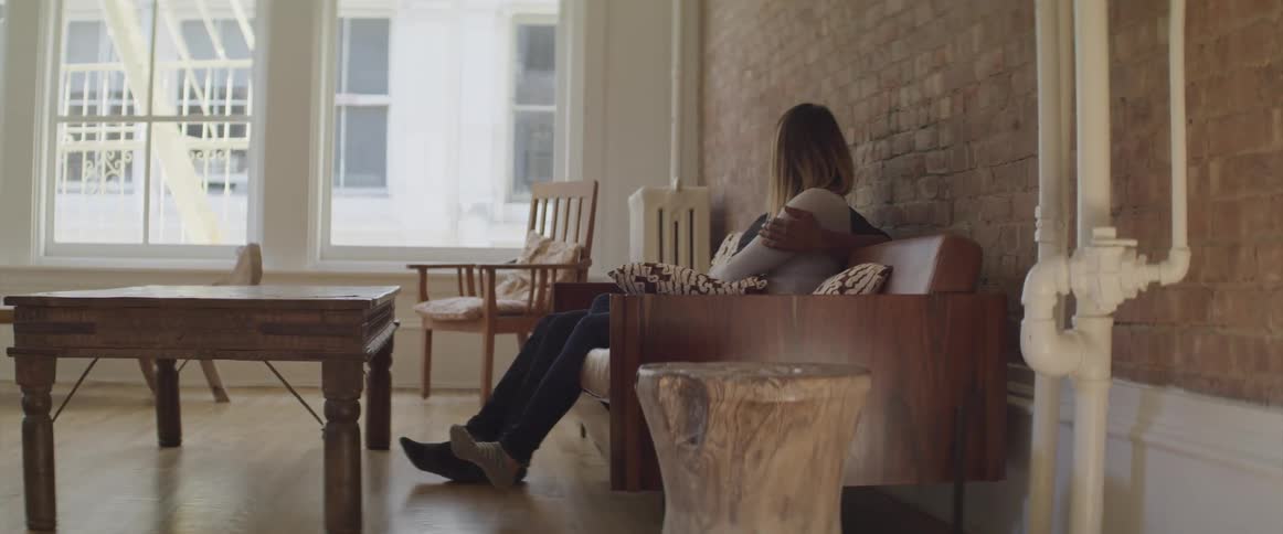A woman sits with her arms crossed on her couch in a sparsely decorated apartment